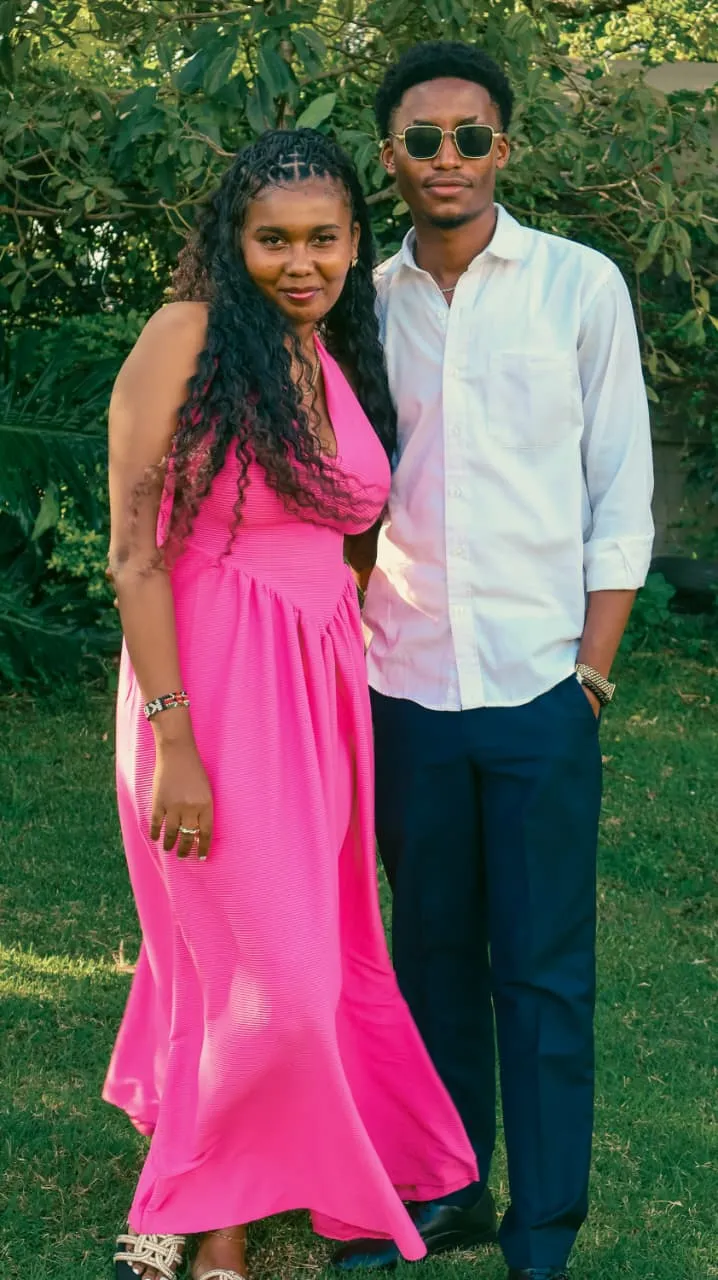 A couple dressed for celebration — she in a pink gown, he in a white shirt — standing close together in golden garden light