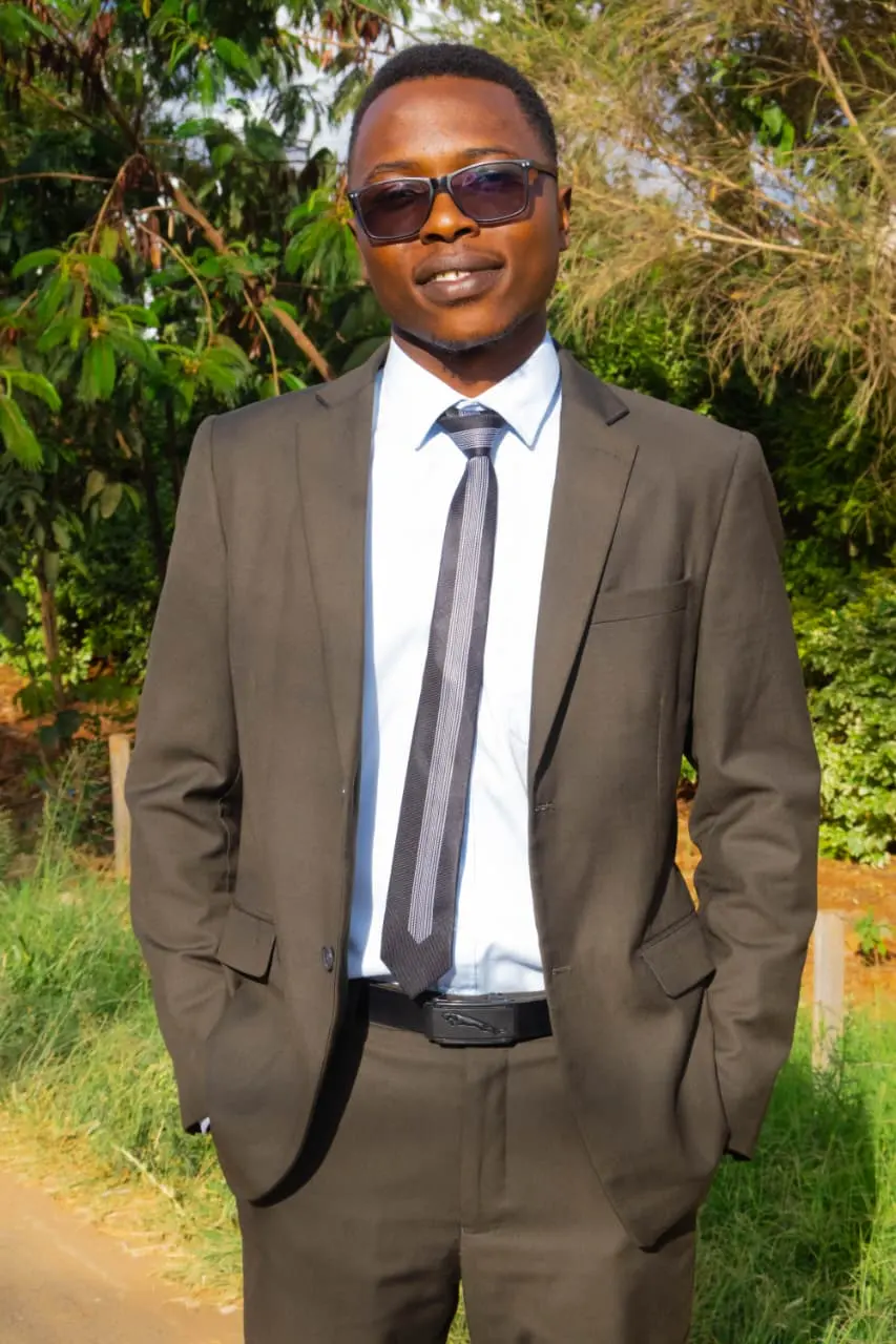 Close portrait of the graduate in his brown suit and sunglasses, hands in pockets, trees behind him